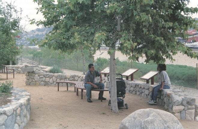 Along a softer section of the Los Angeles River, visitors relax in the shade of a young, native sycamore (Platanus racemosa) in Zanja Madre Park. Interpretive signs behind tell the story of the river.