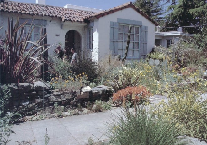 A low, dry-laid stone wall provides a sense of enclosure for the front garden, yet leaves the plantings open for enjoyment by passersby. A tall wooden sculptural element on the left repeats some of the strong lines of New Zealand flax (Phormium), red hot pokers (Kniphofia), a restio (Chondropetalum), and a single columnar cactus (Cereus peruvianus). California poppies (Eschscholzia californica), various aloes, and Phlomis aurea provide springtime flowers in oranges and golds. Photograph by RGT